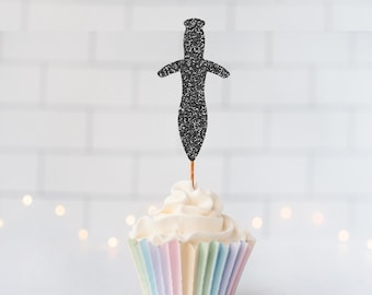 May include: A black glittery dagger-shaped cupcake topper is inserted into a white frosted cupcake. The cupcake sits in a pleated paper liner featuring vertical stripes of pastel colors including light blue, pink, and yellow.