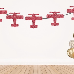 May include: A decorative garland featuring five red glittery airplane cutouts strung together on a thin cord, accompanied by a cluster of gold and gold-glittered balloons positioned to the lower right side of the display.