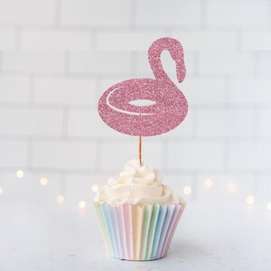 May include: A pink glittery flamingo-shaped cupcake topper attached to a thin wooden pick, inserted into a cupcake with white frosting and a pastel-colored pleated paper liner. The flamingo design features a curved neck and a circular body.