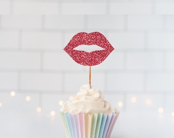 May include: A cupcake with white frosting in a pastel striped paper liner, topped with a red glittery lip-shaped cutout on a thin wooden pick. The decoration features a textured, sparkling surface and is centered above the frosting.
