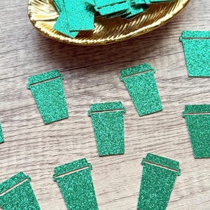 May include: A collection of small, glittery green paper cutouts shaped like coffee cups. Some of the cup-shaped confetti pieces are scattered across the surface, while others are piled inside a gold-coloured, leaf-shaped decorative dish.