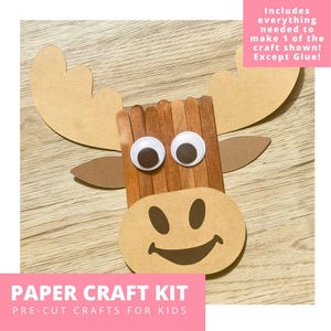 Peut inclure: Un kit de bricolage en papier pour enfants avec un motif d'orignal. L'orignal a des yeux globuleux, des bois en bâtonnets de glace bruns et un visage souriant. Le kit comprend tout le nécessaire pour réaliser un bricolage, sauf la colle. Le texte "PAPER CRAFT KIT" est affiché.
