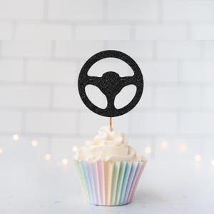 May include: A black glittery steering wheel cake topper attached to a thin wooden pick, inserted into a vanilla cupcake with white frosting in a pastel-coloured pleated paper liner. The topper features a circular design with three spokes.