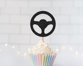 May include: A black glittery steering wheel cake topper attached to a thin wooden pick, inserted into a vanilla cupcake with white frosting in a pastel-colored pleated paper liner. The topper features a circular design with three spokes.