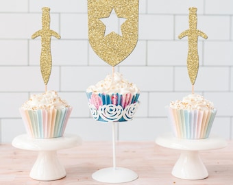 May include: Three cupcakes topped with gold glitter cardstock decorations. Two cupcakes feature sword-shaped toppers, while the center cupcake has a shield-shaped topper with a star cutout. Each cupcake sits in a decorative paper liner.