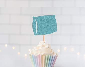 May include: A teal glitter pillow-shaped cupcake topper attached to a thin wooden stick, inserted into a white frosted cupcake with a pastel-colored pleated paper liner. The topper features a textured, sparkling finish on a rectangular pillow shape.