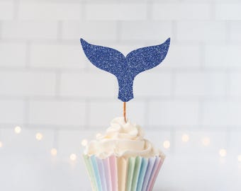 May include: A cupcake topper shaped like a whale tail with a dark blue glitter finish, attached to a thin wooden pick. The topper is inserted into a cupcake with white frosting and a pastel-colored pleated paper liner, creating a decorative accent.