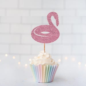 May include: A pink glittery flamingo-shaped pool float cupcake topper attached to a thin wooden pick, inserted into a cupcake with white frosting and a pleated paper liner featuring vertical stripes in pastel shades of pink, blue, and yellow.