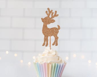 May include: A glittery rose gold reindeer cupcake topper featuring a silhouette of a standing deer with antlers. The topper is inserted into a white frosted cupcake held in a pleated paper liner with vertical stripes of pastel pink, blue, and yellow.