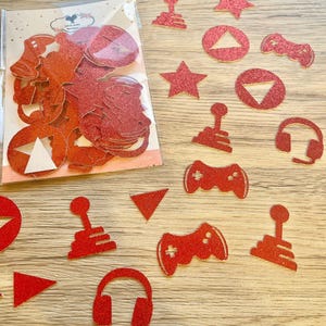 May include: A collection of red glittery paper cutouts featuring gaming-themed shapes including joysticks, game controllers, headsets, stars, and triangles. Some pieces are stored in a clear plastic bag with a label, while others are scattered.