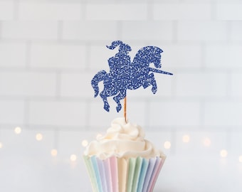 May include: A blue glitter cupcake topper shaped like a knight riding a horse, mounted on a thin wooden pick. The topper is inserted into a cupcake with white frosting and a pastel-striped paper liner, featuring a textured, sparkling finish.