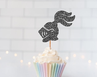 May include: A black glittery knight helmet cupcake topper with a plume, attached to a thin wooden pick, inserted into a white frosted cupcake with a pastel-colored pleated paper liner. The topper features a stylized visor and crest design.