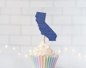 May include: A cupcake topper shaped like the state of California, featuring a dark blue glitter finish. The cutout is attached to a thin wooden pick and is inserted into a cupcake with white frosting and a pastel-colored paper liner.