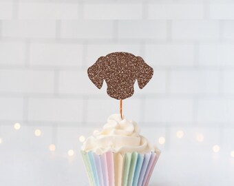 May include: A brown glittery dog head silhouette cupcake topper on a thin wooden pick, inserted into a white frosted cupcake with a pastel-colored pleated paper liner. The dog shape features floppy ears and a rounded snout in a textured finish.