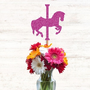 May include: A bright pink glitter carousel horse cutout attached to a wooden stick, positioned above a clear glass vase filled with a vibrant bouquet of fresh gerbera daisies in shades of white, yellow, orange, red, and hot pink.