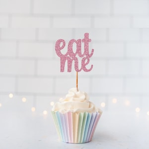 May include: A cupcake with white frosting and a pastel-striped case. A pink glitter "eat me" cake topper is inserted into the cupcake. The background is a white brick wall with soft, out-of-focus lights.