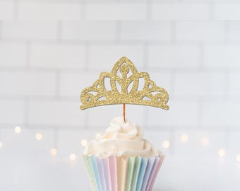May include: A gold glitter tiara cupcake topper sits atop a cupcake with white frosting in a pastel striped paper liner. The tiara features a cutout crown design with a central point and curved side arches, supported by a thin wooden pick.