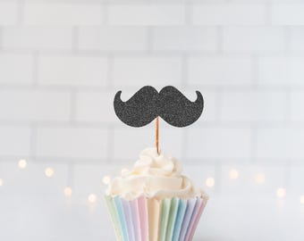 May include: A cupcake topper featuring a black glittery mustache shape attached to a thin wooden pick. The topper is inserted into a cupcake with white frosting and a pleated paper liner with vertical stripes in pastel shades of blue, pink, and yellow.