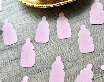 May include: A collection of light pink glittery baby bottle-shaped paper cutouts scattered across a surface, with a larger stack of the same pink glittery baby bottle cutouts resting inside a gold-colored, leaf-shaped decorative dish.