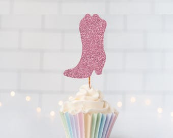 May include: A pink glittery cowboy boot cupcake topper attached to a thin wooden pick, inserted into a cupcake with white frosting and a pastel-colored pleated paper liner. The boot shape is flat and features a textured, sparkling surface.