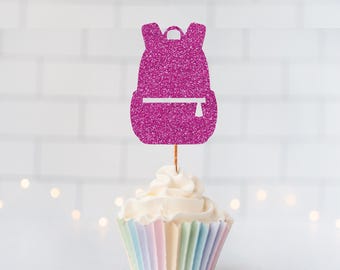 May include: A bright magenta glitter backpack cupcake topper featuring a white horizontal zipper detail. The cutout is attached to a thin wooden pick and is positioned vertically centered in a cupcake with a pastel striped paper liner.