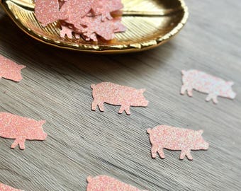 May include: A collection of small, pig-shaped confetti pieces made from light pink glittery material. Several individual pig cutouts are scattered across the surface, while a larger pile of the same pig-shaped confetti rests inside a gold leaf-shaped dish.