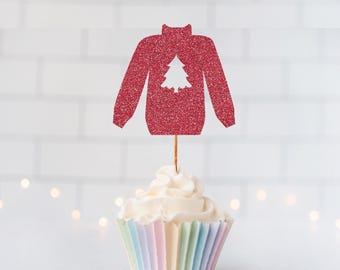 May include: A red glittery sweater-shaped cupcake topper featuring a white cutout of a Christmas tree in the center. The topper is attached to a thin wooden pick and is inserted into a cupcake with white frosting and a pastel striped paper liner.