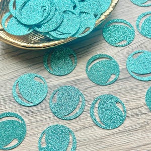 May include: A collection of circular teal glitter confetti pieces, each featuring a small crescent-shaped cutout. Some pieces are clustered inside a gold-toned scalloped bowl, while others are scattered individually across the surface.