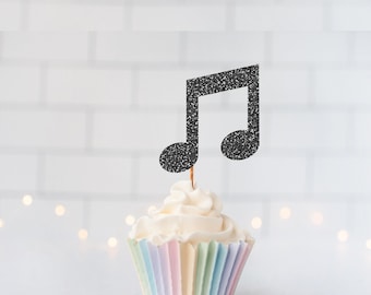 May include: A black glittery musical note cupcake topper on a wooden pick, inserted into a white frosted cupcake with a pastel-colored pleated paper liner. The musical note is shaped like a beamed eighth note with a textured, sparkling finish.