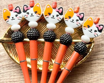 May include: Five orange ballpoint pens featuring a decorative topper shaped like a calico cat face with a pink bow. Each pen includes a white spacer bead, a black textured bead, and a small orange bead at the top, with a silver metal tip.