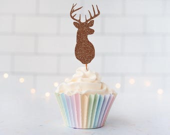 May include: A cupcake topped with a brown glittery deer head silhouette cake topper. The cupcake features white frosting and sits in a pleated paper liner with vertical stripes in pastel shades of pink, yellow, blue, and green.