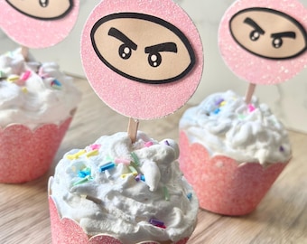 May include: Three cupcakes in pink glittery wrappers topped with white frosting, colorful sprinkles, and a circular pink glittery topper featuring a cartoon face with thick black eyebrows and large eyes on a wooden stick.