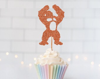 May include: A copper-colored glitter gorilla cupcake topper featuring a silhouette of a gorilla with arms raised, attached to a thin wooden pick, inserted into a white frosted cupcake with a pastel-colored pleated paper liner.