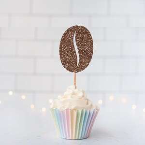 May include: A cupcake topper shaped like a coffee bean with a brown glitter finish, attached to a thin wooden pick. The topper is inserted into a cupcake with white frosting and a pleated paper liner featuring vertical stripes in pastel colors.