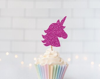 May include: A cupcake topper featuring a glittery fuchsia unicorn head silhouette attached to a thin wooden pick. The cupcake is topped with white frosting and sits in a pleated paper liner with vertical pastel stripes of pink, blue, and yellow.