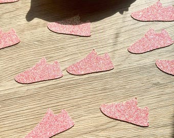 May include: A collection of small, pink glittery paper cutouts shaped like sneakers. The cutouts are scattered across a surface, with a pile of additional matching sneaker-shaped cutouts contained within a gold-colored, leaf-shaped dish.