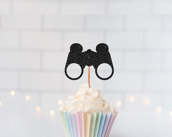 May include: A black glittery binocular-shaped cupcake topper attached to a thin wooden pick, inserted into a white frosted cupcake with a pastel-colored pleated paper liner. The binocular cutout features two circular lenses and a bridge.