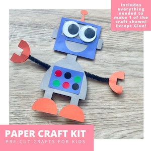 May include: A paper craft kit for kids featuring a colorful robot design. The robot has a blue head with googly eyes, a gray body, orange arms and feet, and a black pipe cleaner. The kit includes everything needed to make one robot, except glue. The text "PAPER CRAFT KIT" is visible.
