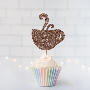 May include: A brown glittery coffee cup cupcake topper featuring a steaming mug silhouette with a handle, mounted on a thin stick and inserted into a white frosted cupcake with a pastel-coloured pleated paper liner for a decorative display.