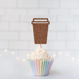 May include: A brown glittery coffee cup topper shaped like a disposable beverage container with a lid. The topper is mounted on a thin wooden pick and is inserted into a cupcake with white frosting and a pastel-colored pleated paper liner.