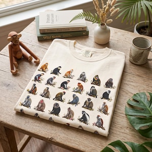 Op de afbeelding: Een crèmekleurig t-shirt met een herhalend rasterpatroon van verschillende apensoorten in diverse houdingen en kleuren. Het shirt heeft een ronde hals en een Comfort Colors merklabel aan de binnenkant van de kraag.