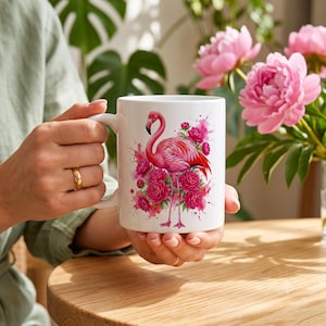 Pode incluir: Uma caneca de cerâmica branca com uma ilustração vibrante de um flamingo cor-de-rosa em pé entre um grupo de flores de peônia rosa brilhante, acentuada com salpicos e manchas de aguarela rosa artística na superfície da caneca.