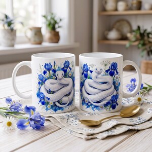 May include: Two white ceramic mugs featuring a detailed illustration of a coiled white snake surrounded by vibrant blue iris flowers and small white daisies. The mugs have a smooth finish, a curved handle, and a clean white interior and exterior.
