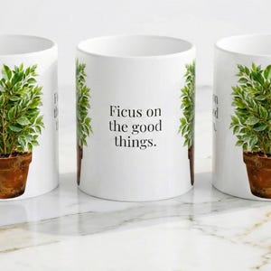 May include: White ceramic mug with a watercolour illustration of a ficus plant in a terracotta pot. The mug has the text "Focus on the good things."
