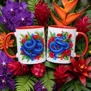 May include: Two white ceramic mugs featuring a vibrant graphic of a coiled blue snake with a red head, surrounded by red tropical flowers and green palm leaves. Each mug has a bright red handle and a matching red interior rim.