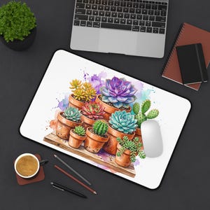May include: A rectangular desk mat featuring a watercolor illustration of various succulents and cacti in terracotta pots arranged on a wooden surface, accented by purple and orange paint splatters on a white background.