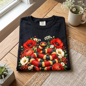 Può includere: Una t-shirt nera con una stampa grafica di un serpente avvolto con squame rosse, gialle e nere, circondato da una composizione di papaveri rossi, margherite bianche e ranuncoli arancioni. L'etichetta interna riporta la scritta Comfort Colors.