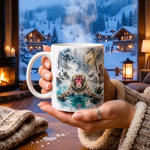 May include: A white ceramic mug featuring a Japanese macaque soaking in a hot spring, with a snowy forest backdrop. The mug is held in front of a window with a view of snow-covered cabins and trees. Steam rises from the mug, suggesting a hot beverage.