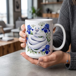 Puede incluir: Una taza de cerámica blanca con una ilustración detallada de una serpiente blanca enroscada rodeada de vibrantes flores de iris azules, pequeñas margaritas blancas y follaje verde. El diseño está centrado en la parte frontal de la taza.