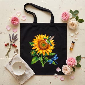 Könnte beinhalten: Eine schwarze Tragetasche mit einem lebendigen Grafikdruck einer großen gelben Sonnenblume, einem grünen und roten Kolibri, der in der Mitte schwebt, und einer leuchtend blauen Libelle, die auf den unteren grünen Blättern ruht, mit zwei schwarzen Tragegriffen.